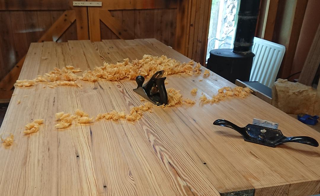 Curled shavings and a plane on a plank table top being planed by hand